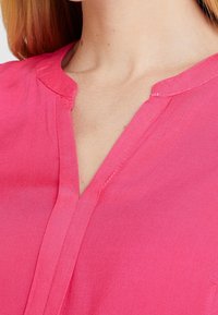 Blouse rose avec un décolleté en V, présentant une texture lisse et un  éclat subtil, mise en valeur par des coutures soignées le long du col et de la patte de boutonnage.