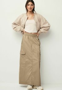 Jachetă bomber bej cu mâneci bufante peste un top crem, asortată cu o fustă maxi lungă, de culoare khaki, cu buzunare tip cargo și un tiv elastic.