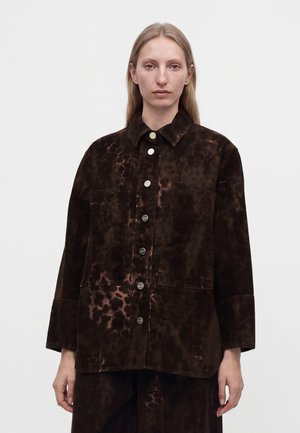 Femme aux cheveux blonds raides porte une veste marron foncé boutonnée avec un motif tie-dye et de grandes poches avant, debout devant un fond uni.