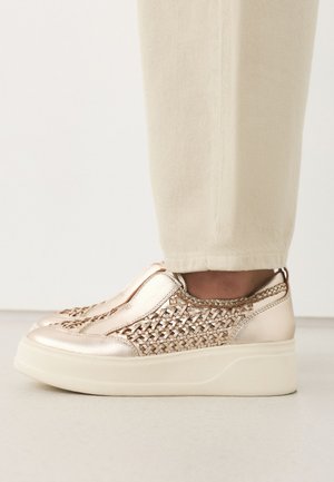 Person trägt goldene Slip-On Plateau-Sneaker mit gewebten Seitendetails und beigen Hosen, steht auf hellem Boden.