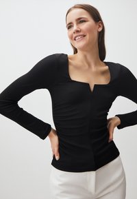 Top nero a maniche lunghe in tessuto a coste con scollatura quadrata e spacco frontale. Presenta dettagli arricciati ai lati. Abbinato a pantaloni di colore chiaro.
