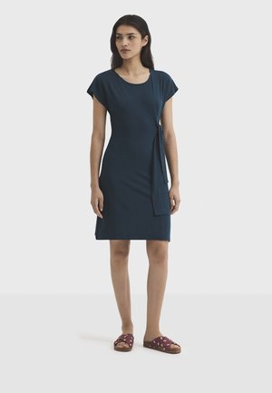 Femme aux longs cheveux noirs portant une robe bleu marine mi-longue avec un détail de nouage sur le côté et des sandales maroon glissées, debout sur un fond blanc.