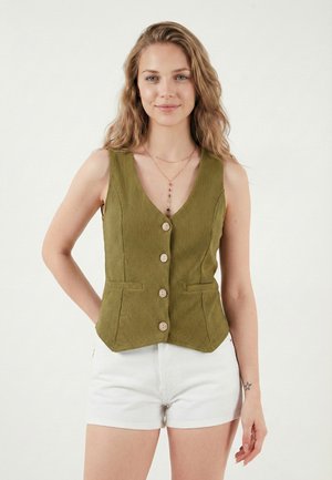 Junge Frau mit welligem Haar, die eine olivgrüne, geknöpfte Weste und weiße Shorts trägt und mit einer Hand hinter dem Rücken steht.