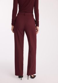 Burgundowe spodnie z wysokim stanem o prostym kroju, gładkiej fakturze i subtelnych detalach w postaci zagnieceń, zestawione z czarnymi butami na obcasie.