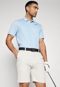 Camisa polo azul clara com um logo preto da Nike, emparelhada com calções brancos e um cinto preto da CK. O sujeito segura um taco de golfe e usa uma luva branca.