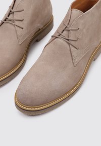 Botas de tobillo de ante beige con puntera redondeada, diseño con cordones y suelas de goma marrón contrastantes con detalles de costura blanca.