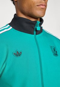 Grønn glidelåsjakke med svart krage; har hvite striper og broderte Adidas- og Liverpool-logoer. Materialet har en glatt tekstur.