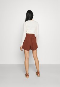 Shorts de couleur rouille avec une taille haute et une ceinture élastique. Associés à un haut à manches longues crème avec des manches texturées. Sandales brunes.