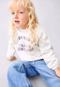 Bambino con lunghi capelli biondi che indossa un felpa grigia chiara e jeans blu, seduto per terra contro uno sfondo semplice.