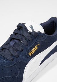 Marineblaue Wildleder-Sneaker mit weißen Akzenten, ausgestattet mit einem goldenen "PUMA"-Logo, schnürbarem Design und strukturiertem Gummilaufsohle.