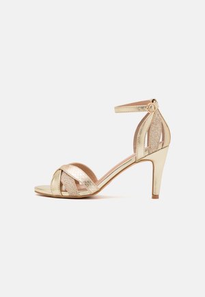 Guld højhælet sandal med åben tå, krydsede remme, ankelrem med spænde og slank mellemhøj hæl på ensfarvet baggrund.