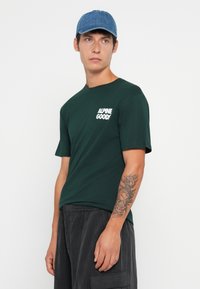 Ung mann med blå caps, mørkegrønn "ALPINE GOODS" t-skjorte og mørke bukser, som viser detaljert tatovering på venstre underarm.