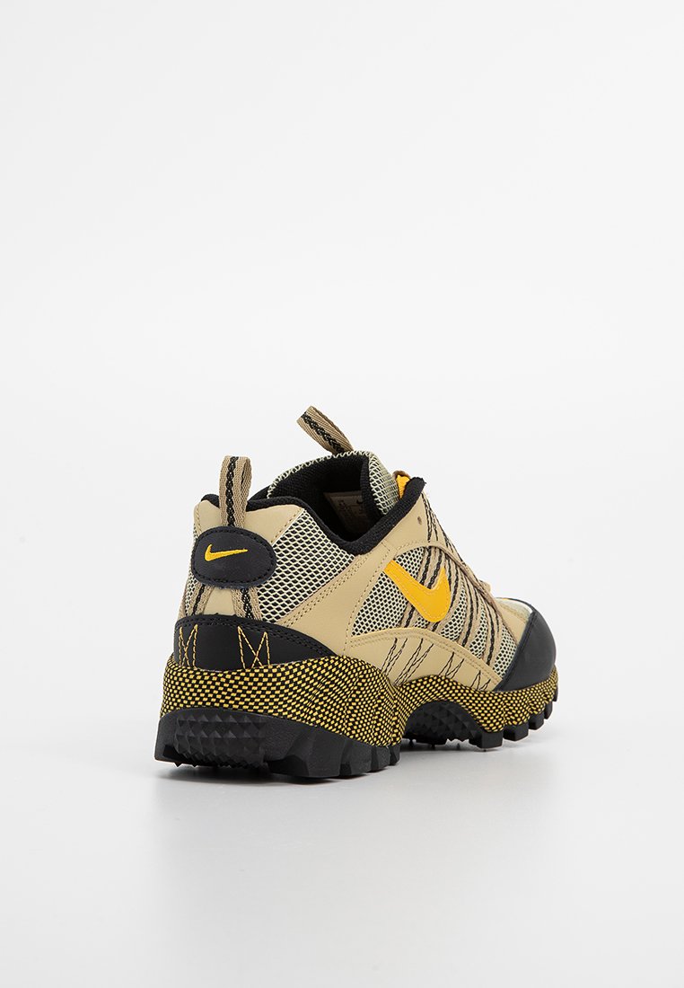 Beżowy but sportowy z czarną podeszwą antypoślizgową, żółtymi akcentami i siateczkowym wierzchem. Posiada wyściełany kołnierz oraz wyraźne logo Nike.