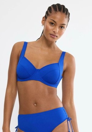 Frau mit geflochtenem Haar trägt einen knallblauen Bikini, steht vor einem schlichten weißen Hintergrund, blickt geradeaus mit einem neutralen Ausdruck.