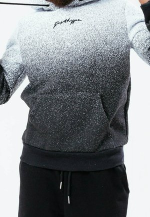 Mies, jolla on mustavalkoinen pilkullinen huppari, jossa on etutasku ja kiristysnauhat, yhdistettynä mustiin housuihin, kädet kohotettuina.