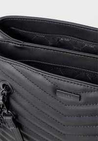 Czarna torebka z sztucznej skóry o pikowanym wzorze chevron, z zamykaną na zamek kieszenią wewnętrzną i metalowym logo. Posiada łańcuszkowy pasek.