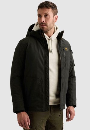 Homme aux cheveux courts portant une veste noire à capuche par-dessus un sweat à capuche gris clair et un pantalon vert olive, regardant sur le côté.