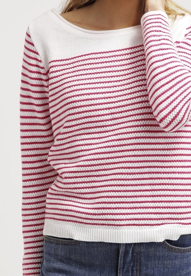 Femme portant un pull en tricot blanc avec des rayures horizontales roses et un jean bleu, main touchant les cheveux près du visage, torse visible.
