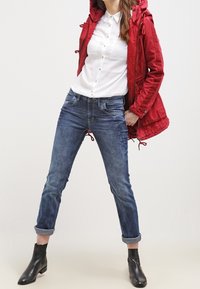 Rode hoodie met verstelbare trekkoord, witte overhemden, en blauwe jeans met opgerolde manchetten. Afgewerkt met zwarte enkellaarsjes.