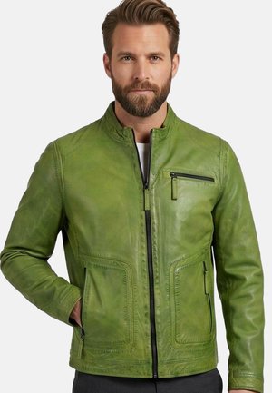 Mann mit Bart, der eine grüne Lederjacke mit Reißverschluss und Reißverschluss- sowie aufgesetzten Taschen trägt, steht mit einer Hand in der Tasche vor weißem Hintergrund.