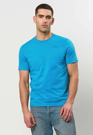 Mann mit kurzen dunklen Haaren, der ein hellblaues kurzärmeliges T-Shirt und hellblaue Jeans vor einem einfarbigen Hintergrund trägt.