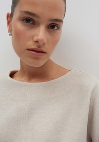 Indumento beige chiaro, testurizzato, con un ampio scollo rotondo. La modella ha un trucco neutro e orecchini a bottone argento. Sfondo uniforme.