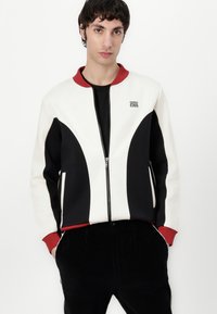 Crewneck-jakke i hvit, svart og rød. Har en dristig, buet paneldesign, frontglidelås og sidelommer. Laget av teksturert stoff.