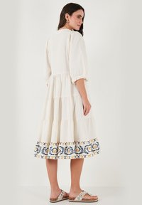 Tiered white dress with elbow-length sleeves and embroidered blue and yellow floral pattern at the hem. Model wearing sandals.