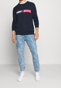 Granatowy sweter z dzianiny z logo „TOMMY JEANS”, zestawiony z jasnoniebieskimi dżinsami z wzorzystą grafiką oraz białymi trampkami.