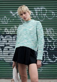 Πλεκτό πουλόβερ μέντας με ριμπ λαιμόκοψη, συνδυασμένο με μαύρη φούστα με πιέτες. Το μοντέλο κρατά ένα λευκό λούτρινο ζωάκι. Φόντο με γκράφιτι.