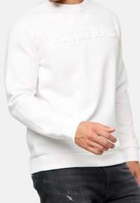 Hvid sweatshirt lavet af blødt stof, med oprebne bogstaver "INDICODE JEANS" på forsiden, samt ribbede manchetter og kant. Casual pasform.