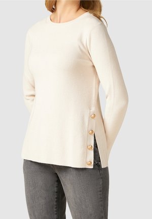 Vrouw draagt een crèmekleurige geribbelde trui met gouden knopen aan de zijsplit, gecombineerd met donkergrijze jeans, tegen een effen achtergrond.