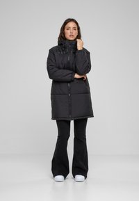 Abrigo acolchado negro con cuello alto, cierre frontal con cremallera y mangas cortas abullonadas. Acentos texturizados y un diseño hasta la rodilla.