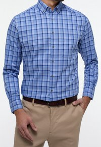 Blauwe geruite overhemden met verschillende tinten blauw en wit, voorzien van een puntige kraag en bruine knopen, gedragen met beige broeken.