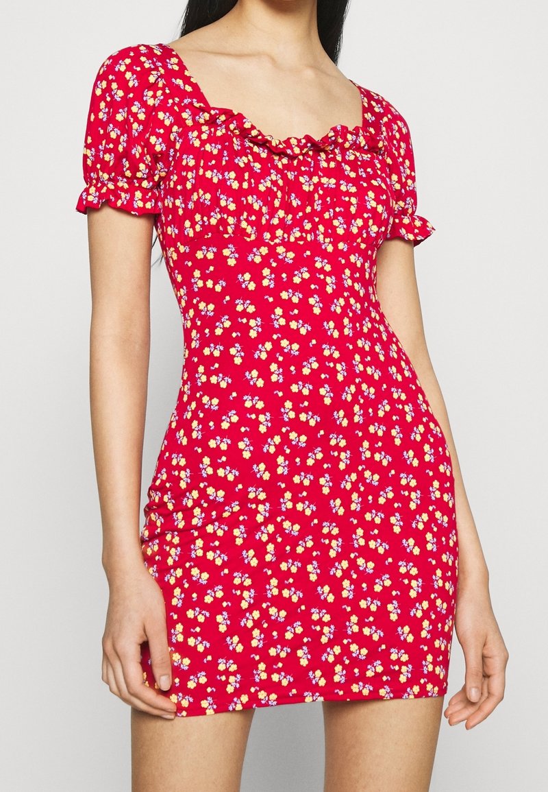 Robe rouge ajustée à manches courtes avec un petit imprimé floral jaune et violet et un col volanté sur une femme debout.