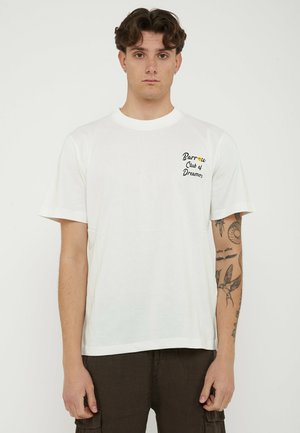 Jeune personne aux cheveux courts et bouclés portant un t-shirt blanc avec le texte « Barrow Club of Dreamers » et des tatouages sur le bras gauche.