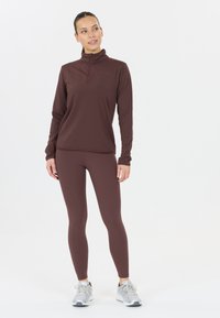 Burgundy pitkähihaisen paita, jossa on puolivetoketju kaulus, yhdistettynä sopiviin legginsseihin; mallilla on päällä harmaat urheilukengät. Sujuva, istuva materiaali.