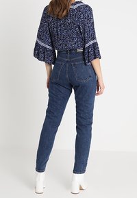 Džíny s vysokým pasem v modré denimové barvě s přiléhavým střihem, s jednou zadní kapsou, nošeny s bílými kotníkovými botami. Texturovaná látka a klasický střih.