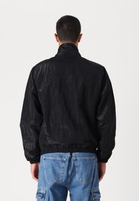 Jachetă neagră tip windbreaker cu o textură lucioasă, talie elastică și logo-ul „LACOSTE” embosat pe spate. Purtați cu blugi cargo albaștri.