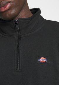 Black zip-up sweatshirt with a high collar, featuring a textured fabric and a multicolored Dickies logo patch on the chest.