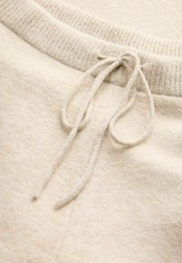 Primo piano di un tessuto a maglia beige con cintura a coste e cordino annodato su un materiale morbido e testurizzato.