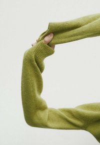 Vihreä neulepuseron hiha, jossa on ribbikuvio ja pitkitetty muoto, pidetään kädessä, joka osoittaa joustavuutta ja pehmeää materiaalia.