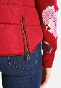 Gros plan d'une personne portant un jean bleu, un pull rouge à manches longues avec motif floral, et une veste matelassée rouge avec une fermeture éclair verticale dorée et un tirette en forme de lettre S.