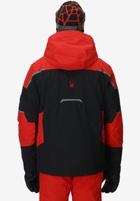 Persona che indossa un giacca invernale nera e rossa, pantaloni da neve abbinati, guanti neri e un berretto a righe nere e rosse, girata di spalle su sfondo bianco.