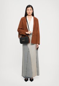 Frau trägt eine braune fransige Jacke, ein weißes Oberteil, einen Jeans-Maxikleid mit Patchwork, schwarze spitze Schuhe und hält eine schwarze Love Moschino Umhängetasche.