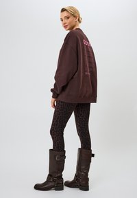 Sudadera marrón de gran tamaño con texto en rosa "GOOD KARMA" en la parte posterior, combinada con leggings de leopardo negros y botas marrones hasta la rodilla con hebillas.