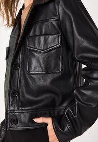 Veste en cuir noire à coupe décontractée, avec deux poches poitrine, un col pointu et une fermeture à boutons. La texture est lisse et brillante.