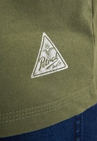 Olive green cotton fabric with ribbed texture; features a white triangular logo patch at the bottom right. Visible stitching detail.