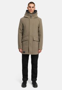 Olivgrön, huvad vinterjacka med dragkedja fram, två stora fickor och en texturerad tyg. Matchas med svarta byxor och stövlar.