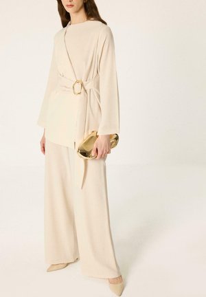 Donna indossa un top avvolgente crema con fibbia dorata, pantaloni a gamba larga, tacchi a punta, tiene una clutch dorata lucida su uno sfondo semplice.